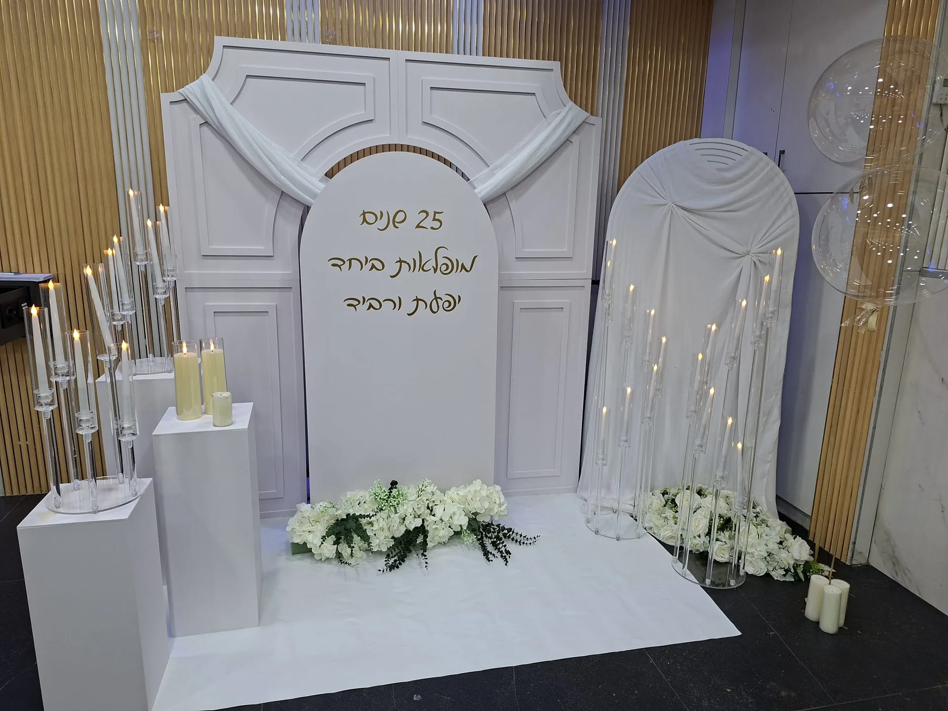 תפאורה לבנה עם קשתות, נרות, פרחים לבנים ובלונים שקופים לצד שלט לציון 25 שנים